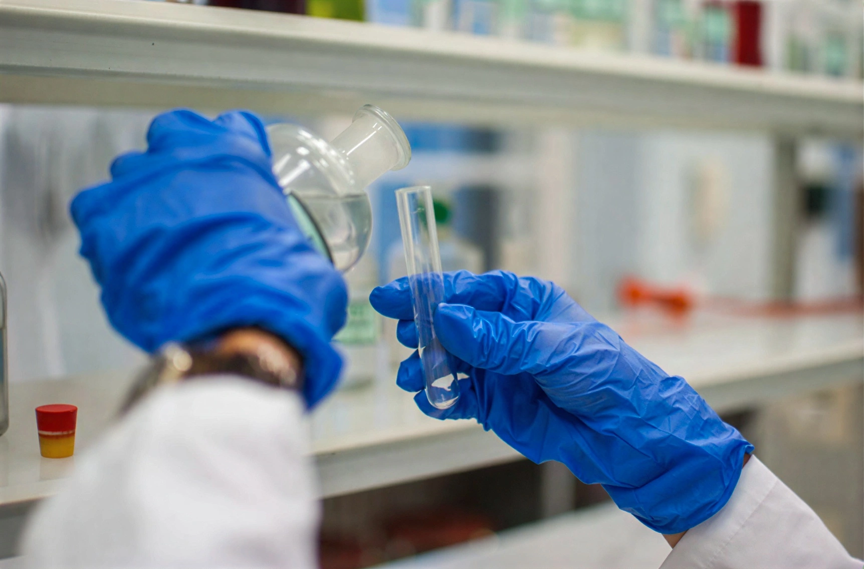 A close-up of a person in a white lab coat and bright blue nitrile gloves carefully pouring a clear liquid from a glass reagent bottle into a test tube.